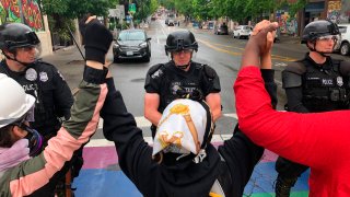 Protesters stand holding up their arms in front of a road blocked by police in the Capitol Hill Organized Protest zone early Wednesday, July 1, 2020, in Seattle. Police in Seattle have torn down demonstrators' tents in the city's so-called occupied protest zone after the mayor ordered it cleared.
