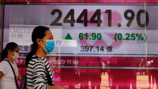 In this April 20, 2020, file photo, people wearing face masks walk past a bank electronic board showing the Hong Kong share index at Hong Kong Stock Exchange.