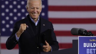 Joe Biden smiles while holding up a hand in front of an American flag.