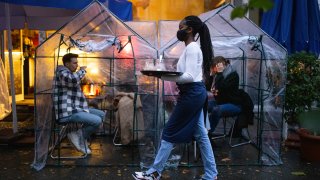 People sitting inside plastic tents at an outdoor cafe with a waitress wearing a face mask carrying a serving tray.