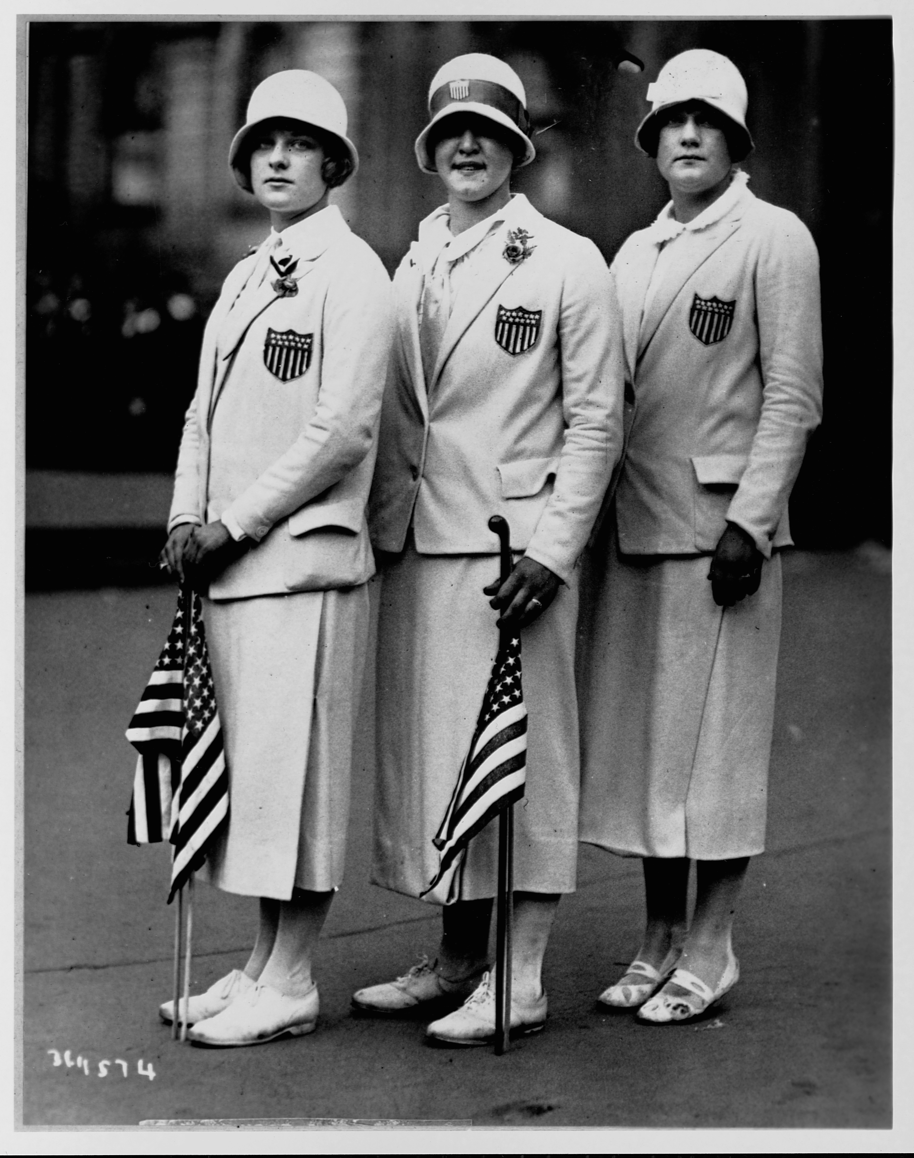 Aileen Riggin, Gertrude Ederle, and Helen Wainright stand in their Olympic team uniforms and hold American flags, after returning from the 1920 Olympics in Antwerp, Belgium.