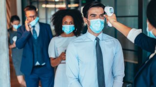 Business colleagues with protective face masks using infrared thermometer for measuring temperature before entrance in office.