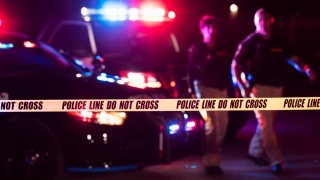 Two multi-ethnic police officers standing in front of patrol cars, behind police tape, wearing bulletproof vests and duty belts. The policewoman is a mature African-American woman in her 40s. Her partner is a mid adult man in his 30s. It is nighttime and the emergency lights on top of the vehicles are flashing. The focus is on the cordon tape in the foreground.