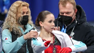 Figure skating coaches Eteri Tutberidze (L) and Sergei Dudakov (R) help ROC athlete Kamila Valieva to put on a sweat jacket