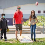 two kids and their teacher stand outside with a cornhole board between them while a bean bag drops from the sky