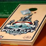 a bean bag slides up a cornhole board with the "port city baggers" logo that features a ship, an anchor, and a pirate throwing a bean bag.