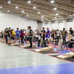 long line of cornhole boards inside a gymnasium, with players lined up throwing bean bags