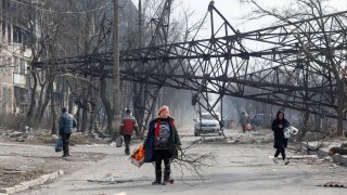 Local residents walk near a fallen electricity pylon and an apartment building destroyed in the course of Ukraine-Russia conflict in the besieged southern port city of Mariupol, Ukraine March 25, 2022.