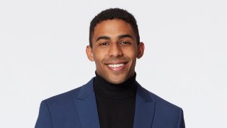 A publicity image of Joshua Tylerbest from ABC's "The Bachelorette." Tylerbest is smiling against a white backdrop, wearing a blue blazer and black shirt.