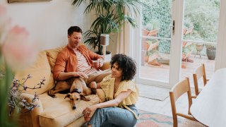 A beautiful young couple relax together in a stylish living room. The male sits on a soft, luxurious velvet yellow sofa as a lurcher dog lies curled up beside him. The female sits on a rug on the ground in front of her partner. They smile.