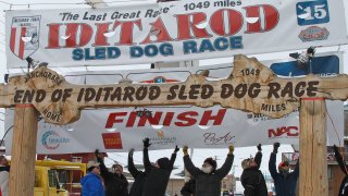 FILE – Volunteers help raise the Iditarod finishers banner at the burled arch finish line in Nome, Alaska, March 16, 2015.