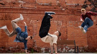 Freeze frame of all male breakdancing team jumping in air against brick wall outdoors