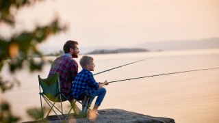 Side view portrait of father and son sitting together on rocks fishing with rods in calm lake waters with landscape of setting sun, both wearing checkered shirts, shot from behind tree