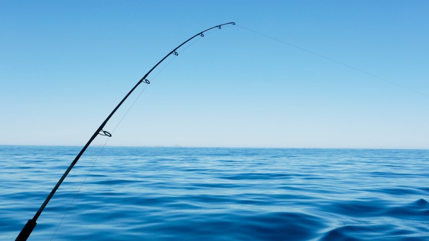 File. Fishing rod bending while catching a fish. Deep blue ocean in the background. Blue sky and copy space.