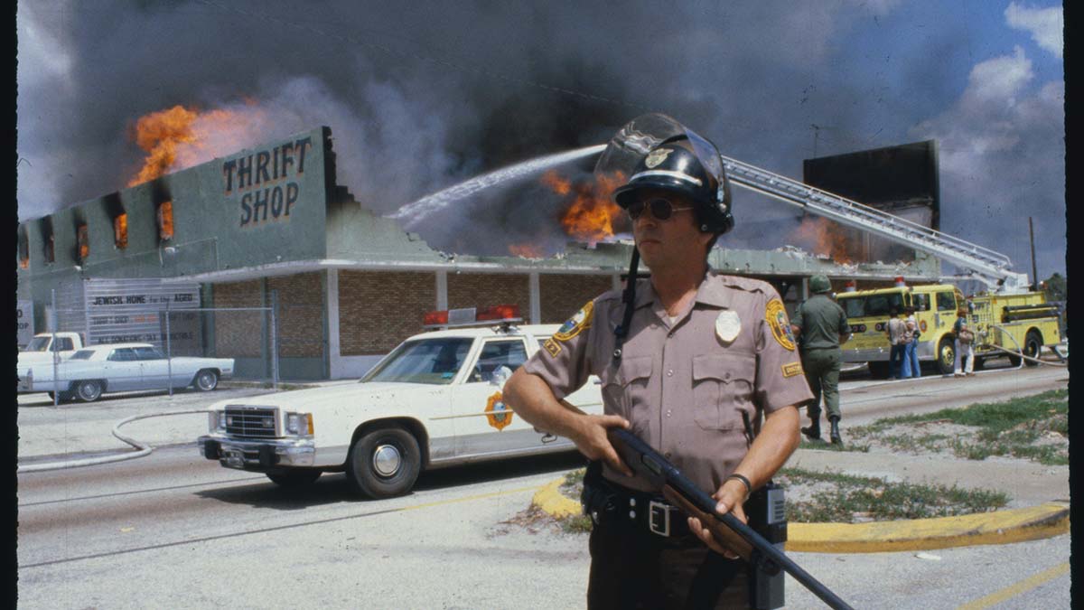(Original Caption) Miami: With a Dade County policeman standing guard, firemen battle a blaze at a thrift shop during the third day of racial violence. The fireman’s helmet is red, the flames are orange, and the gunstock is brown.