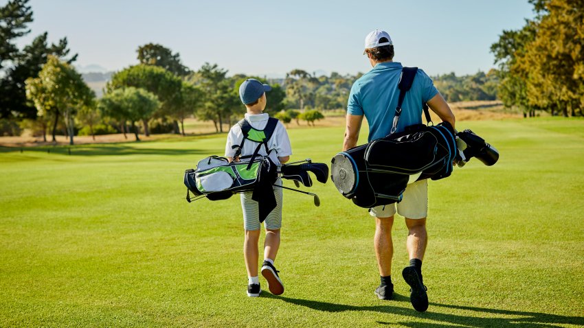 Rear view of man walking with boy on golf course. Father and son are carrying bags at sports field. They are in sportswear.