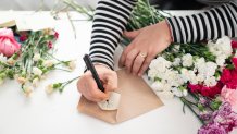 Close up photo of mature woman hands arranging carnation flower bouquet in living room for selling online. She is sitting on sofa next to window, writing a message on envelope and having video call on digital tablet. Shot under daylight.