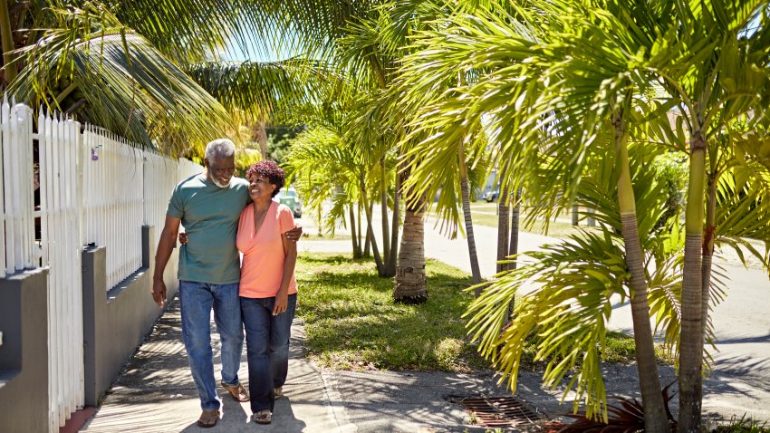 Full length view of Black couple in their 70s wearing casual clothing and approaching camera with arms around each other, talking, and laughing.